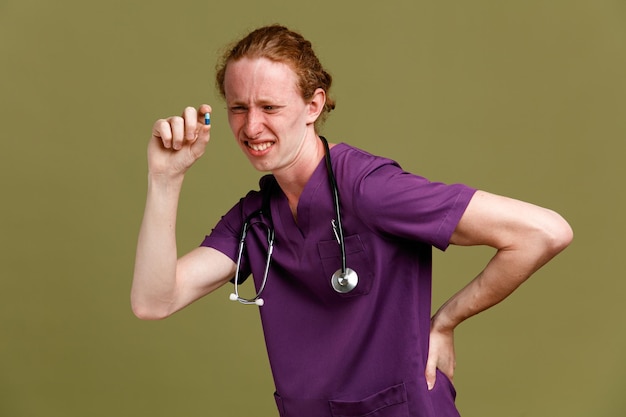 Infeliz poniendo la mano en la espalda sosteniendo pastillas joven médico masculino vistiendo uniforme con estetoscopio aislado sobre fondo verde