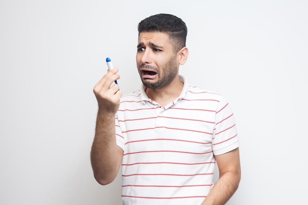 Infeliz preocupação doente jovem adulto em t-shirt branca em pé e segurando e olhando para o termômetro são chocados por alta temperatura. interior, isolado, fundo branco, tiro de estúdio