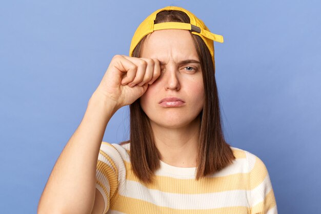 Infeliz, triste, disgustada adolescente con camiseta a rayas y gorra de béisbol, aislada sobre fondo azul, sintiendo fatiga frotándose los ojos cansada y exhausta