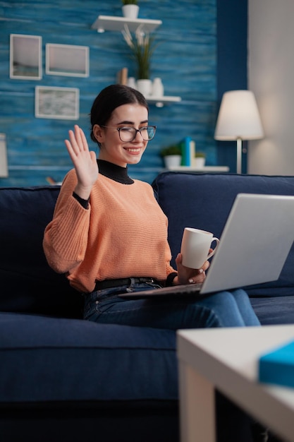 Foto influenciador con anteojos sentado en el sofá sosteniendo una taza y una computadora portátil en el regazo. estudiante sonriente en el sofá saludando en conferencia de videollamada. programador trabajando a distancia desde la oficina en casa en la sala de estar.