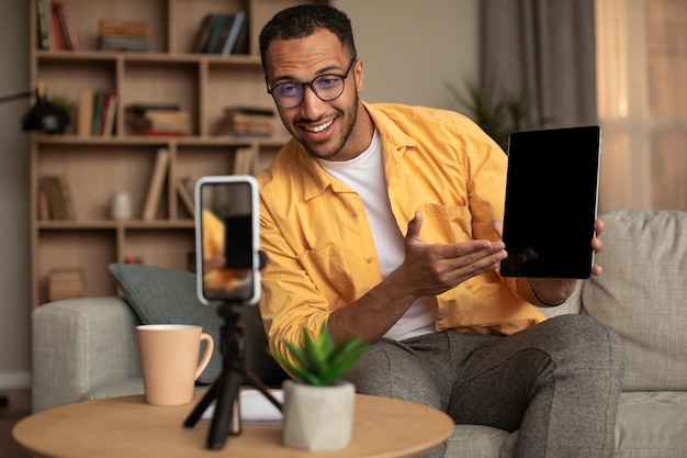 Influenciador masculino negro animado filmando revisão de vídeo do novo tablet digital no smartphone em casa