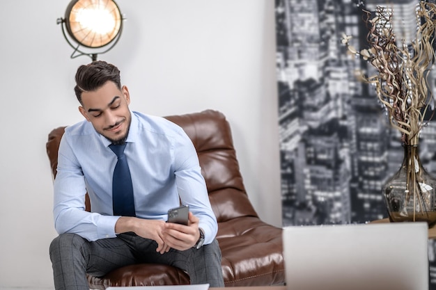 Informação importante. Feliz jovem elegante de gravata sorrindo olhando para smartphone sentado na poltrona de couro