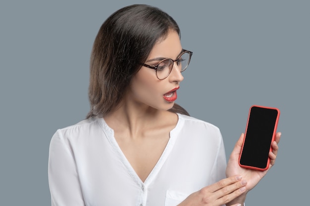 Información útil. Grave mujer exitosa con cabello largo oscuro con gafas en blusa blanca mirando smartphone diciendo