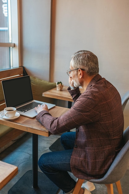 Information. Erwachsener Mann in eleganter Jacke schaut auf den Laptop-Bildschirm, während er tagsüber mit Kaffee drinnen am Tisch sitzt