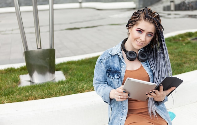 Informelle Studentin mit Afro-Zöpfen und hellem Make-up mit Tablet im Freien