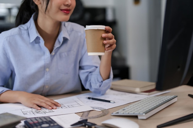 Ingeniera arquitecta asiática mujeres leyendo el plan de información de arquitectura en la computadora y bebiendo chocolate caliente mientras trabajan para comprobar el dibujo del edificio y el plan de construcción en el plano