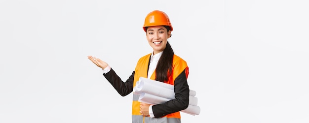 Una ingeniera arquitecta asiática profesional sonriente con casco de seguridad presenta un proyecto de construcción que señala con la mano a la izquierda mientras lleva un plano dando un discurso presenta un gráfico o un plan de construcción