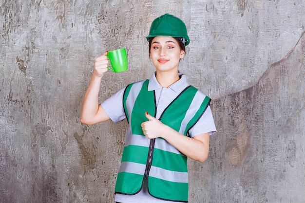 Ingeniera en casco verde sosteniendo una taza de café verde y mostrando signo de disfrute.