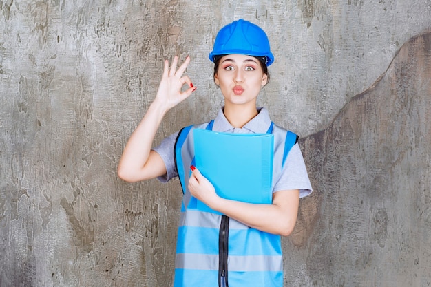 Ingeniera en uniforme azul y casco sosteniendo una carpeta de informe azul.