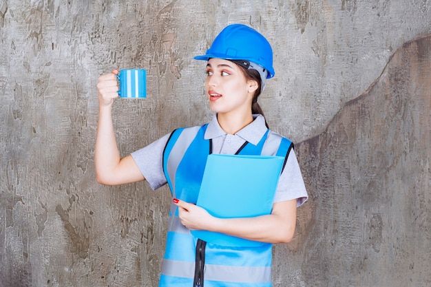 Foto ingeniera en uniforme azul y casco sosteniendo una taza de té azul y una carpeta de informe azul.