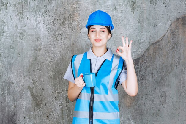 Ingeniera en uniforme azul y casco sosteniendo una taza de té azul y mostrando signo de disfrute.