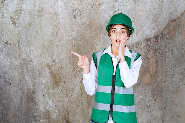 Ingeniera en uniforme verde y casco de pie en la pared de hormigón y apuntando hacia el lado izquierdo