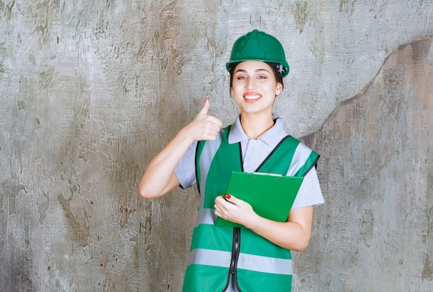 Ingeniera en uniforme verde y casco sosteniendo la carpeta del proyecto y mostrando un signo de mano positivo