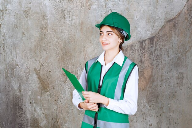 Ingeniera en uniforme verde y casco sosteniendo una carpeta de proyecto verde.