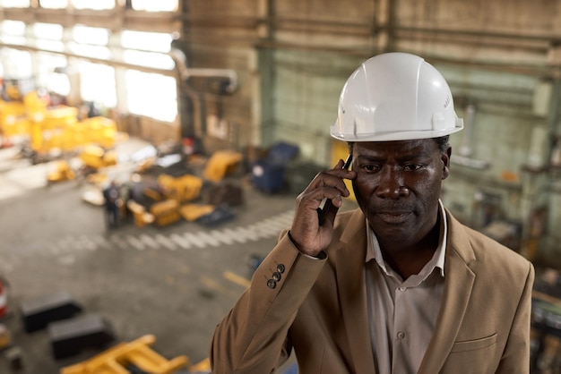 Ingeniero africano con casco de trabajo que se ve muy serio mientras habla por teléfono móvil con un compañero parado en la planta