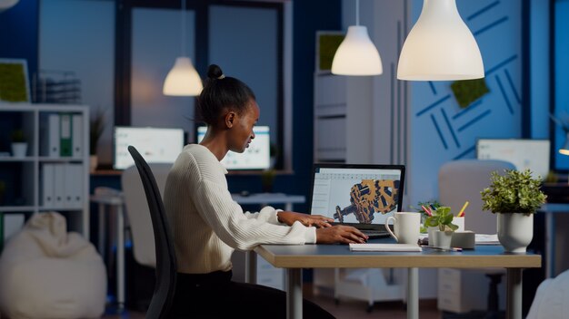 Ingeniero africano trabajando a altas horas de la noche en un modelo 3d de engranajes industriales en la computadora portátil haciendo horas extraordinarias en la oficina de puesta en marcha. freelancer estudia la idea del prototipo en la pc que muestra el software cad en la pantalla del dispositivo