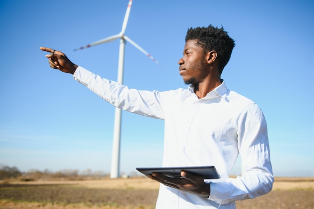 Ingeniero africano vistiendo casco blanco de pie con tableta digital contra la turbina eólica en un día soleado