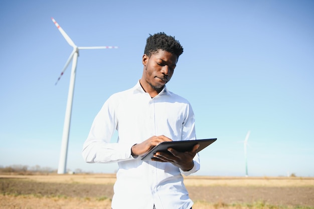 Ingeniero africano vistiendo casco blanco de pie con tableta digital contra turbina eólica