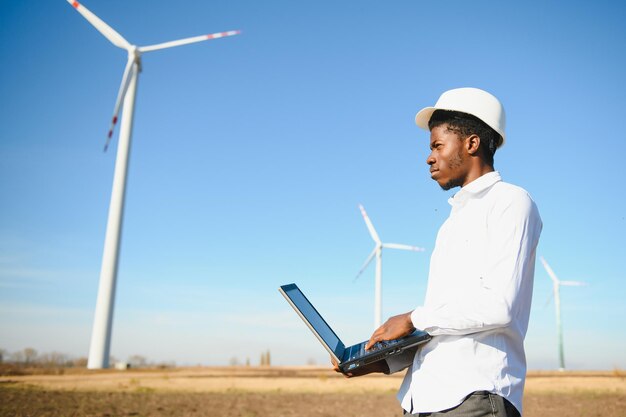 Ingeniero africano vistiendo casco blanco de pie con tableta digital contra turbina eólica