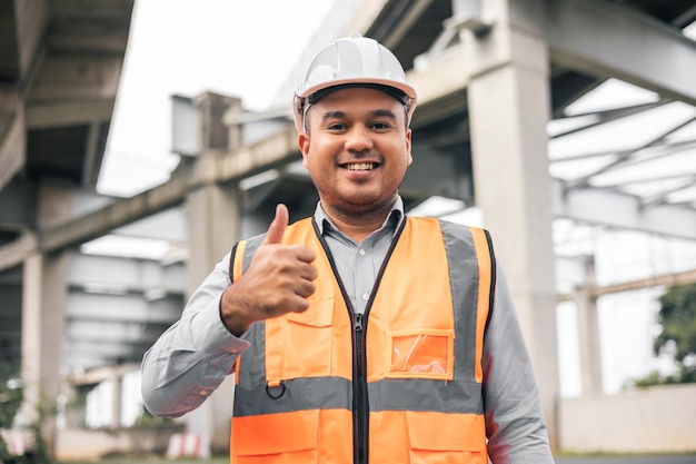 Ingeniero asiático guapo o arquitecto que mira la construcción con casco de seguridad blanco en el sitio de construcción De pie en el sitio de la carretera de hormigón Mostrando los pulgares hacia arriba