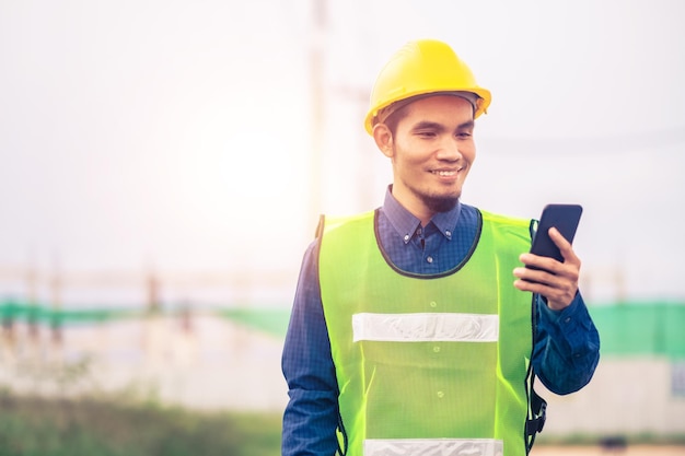 Ingeniero asiático sosteniendo un teléfono inteligente parado al aire libre en la construcción del sitio publicando un feliz éxito en la construcción del ingeniero con la planificación de objetivos