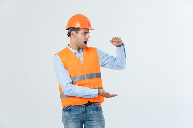 Ingeniero de barba feliz sosteniendo la mano a un lado y explicando algo, chico vestido con camisa caro y jeans con chaleco amarillo y casco naranja, aislado sobre fondo blanco.