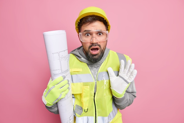 El ingeniero barbudo asustado desconcertado sostiene que el plano arquitectónico viene en el sitio de construcción por miedo a que algo use chaleco reflectante de casco y poses de guantes contra la pared rosada del estudio.