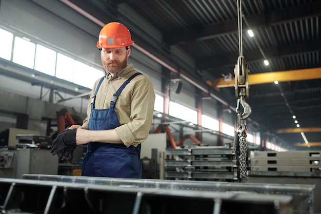 Ingeniero barbudo en ropa de trabajo y casco de pie junto a un enorme detalle de hierro mientras controla el proceso de trabajo en la fábrica.