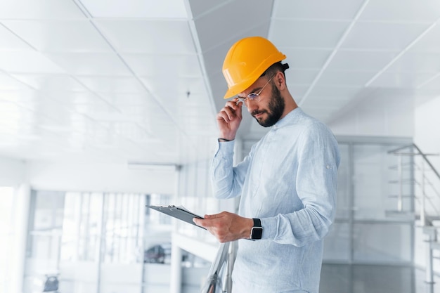 Ingeniero de bloc de notas con ropa blanca y casco protector naranja de pie y trabajando en interiores