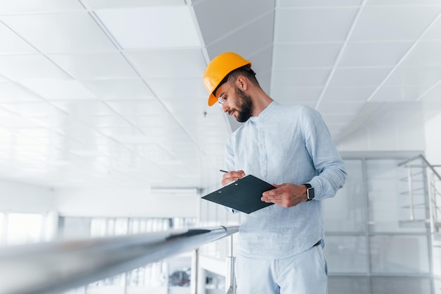 Ingeniero de bloc de notas con ropa blanca y casco protector naranja de pie y trabajando en interiores