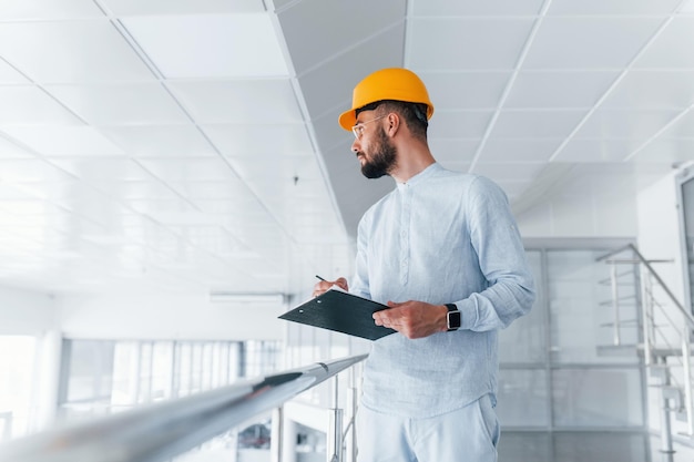 Ingeniero de bloc de notas con ropa blanca y casco protector naranja de pie y trabajando en interiores