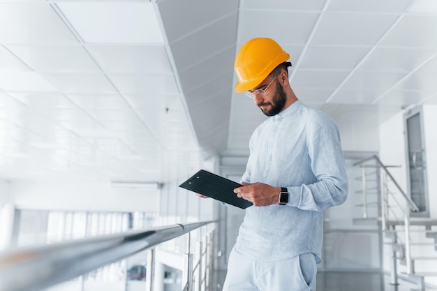 Ingeniero de bloc de notas con ropa blanca y casco protector naranja de pie y trabajando en interiores