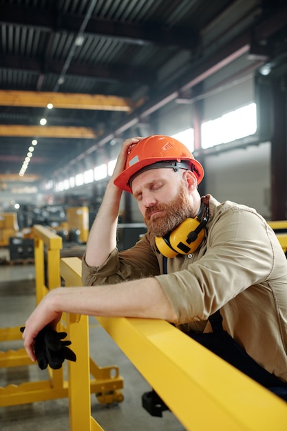 Foto ingeniero cansado o enfermo en ropa de trabajo y casco apoyado en la barra mientras se toma un descanso en medio de la jornada laboral en la fábrica.