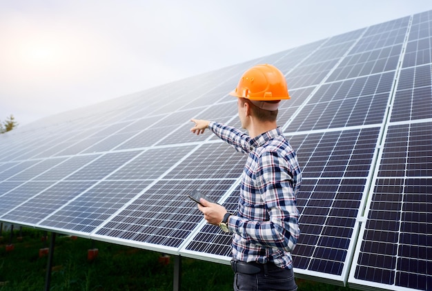 Ingeniero en casco con tableta en manos se encuentra cerca de paneles solares en plantación verde