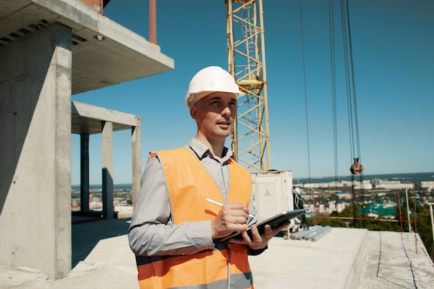Un ingeniero con un chaleco naranja y un casco de control de construcción blanco realiza una inspección con una tableta en sus manos contra el de un sitio de construcción y una grúa torre