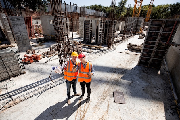 El ingeniero civil y el arquitecto vestidos con chalecos de trabajo naranjas y murciélagos duros discuten el proceso de construcción en el sitio de construcción abierto con material de construcción. .