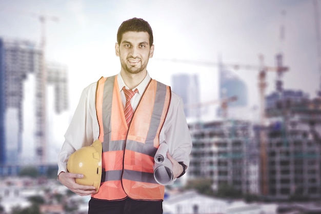 Ingeniero civil hispano sosteniendo un plano y un casco sonriendo con experiencia en construcción