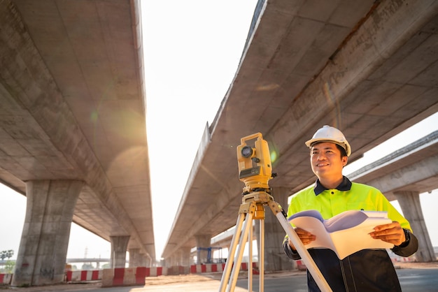 Un ingeniero civil o agrimensor de pie sosteniendo un plano con una sonrisa feliz en el sitio de construcción de carreteras Equipo de agrimensor Carretera