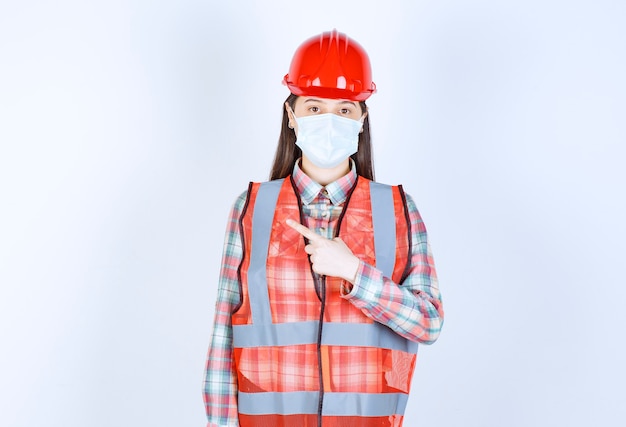 Ingeniero de construcción femenino en casco rojo y máscara de seguridad apuntando hacia la izquierda.