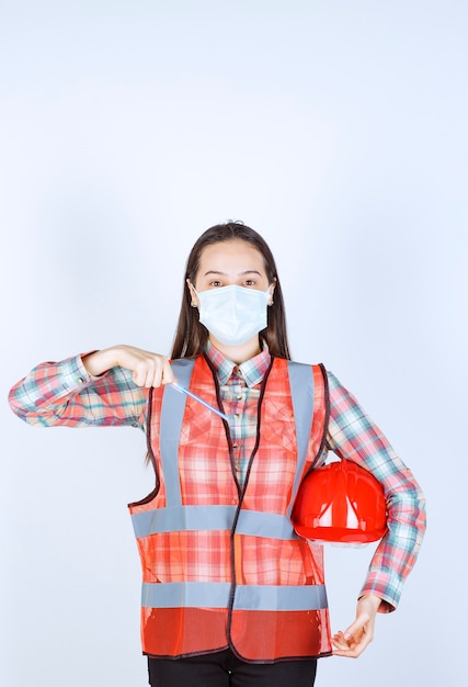 Foto ingeniero de construcción femenino en máscara de seguridad y un casco rojo debajo de sus brazos sosteniendo una pluma