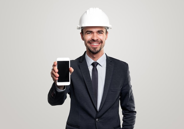 Ingeniero de construcción masculino barbudo adulto feliz en traje formal y casco demostrando teléfono móvil con pantalla negra en blanco sobre fondo gris