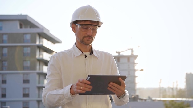Un ingeniero de construcción o arquitecto que lleva una camisa blanca y un casco está usando una tableta digital mientras inspecciona un sitio de construcción en la vista frontal del atardecer.