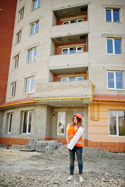 Foto ingeniero constructor mujer en chaleco uniforme y casco protector naranja mantenga rollo de papel de dibujo de negocios contra el nuevo edificio