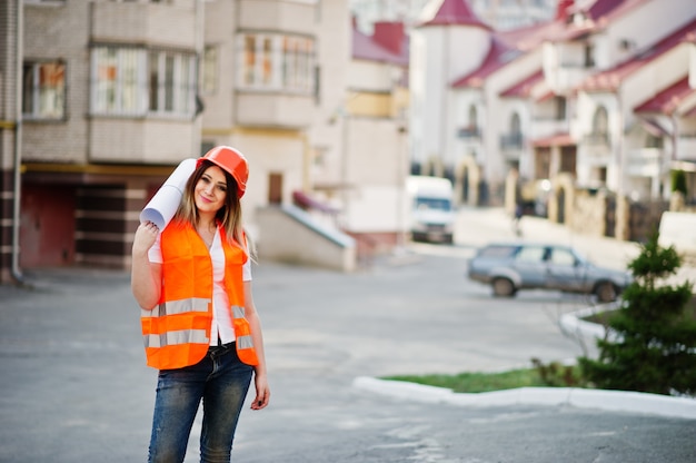 Ingeniero constructor mujer en chaleco uniforme y casco protector naranja mantenga rollo de papel de dibujo de negocios contra el nuevo edificio