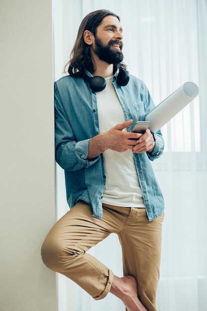 Ingeniero contento de pie con su teléfono inteligente gráfico y moderno junto a la pared y mirando hacia otro lado con una sonrisa