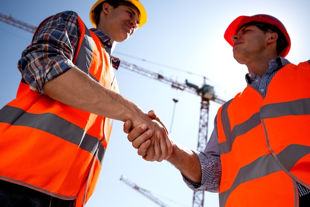 El ingeniero estructural y el arquitecto vestidos con chalecos de trabajo naranja y cascos se dan la mano en el sitio de construcción cerca de la grúa. .