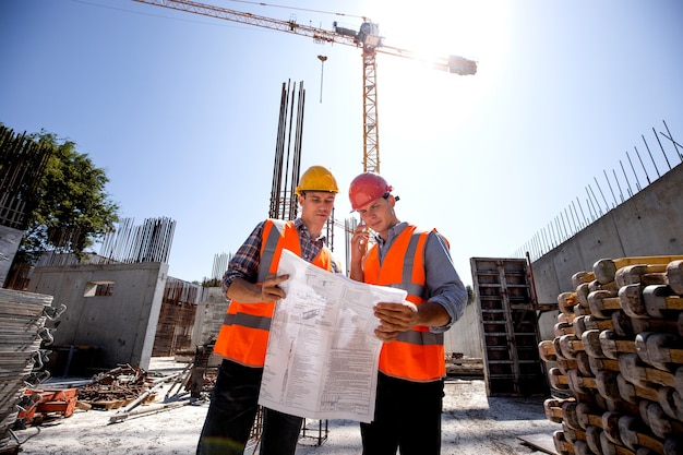 El ingeniero estructural y el gerente de construcción con chalecos de trabajo naranjas y cascos duros discuten la documentación y hablan por teléfono en el sitio de construcción al aire libre junto a la grúa. .