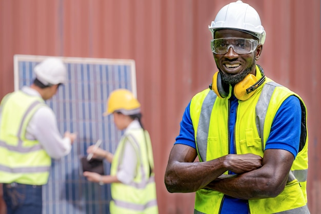 Ingeniero de fábrica hombre africano confianza permanente con vestido verde suite de trabajo y casco de seguridad. El personal de los trabajadores que controla el panel de células solares para la construcción de energía renovable. trabajador de la industria en funcionamiento.