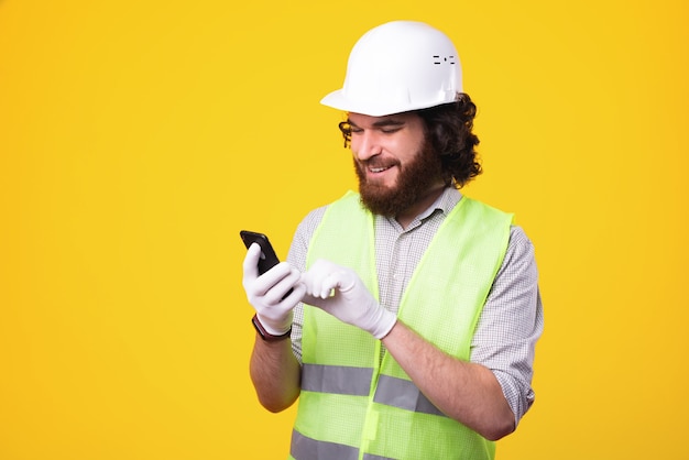 Un ingeniero feliz está enviando mensajes de texto a alguien a través de su teléfono cerca de una pared amarilla