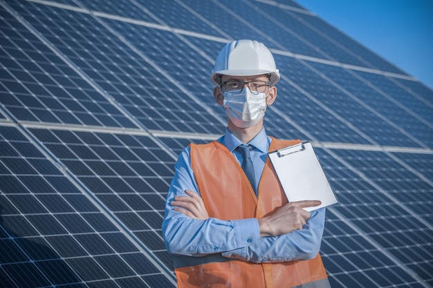 Ingeniero, hombre de uniforme y máscara, gafas de casco y chaqueta de trabajo sobre un fondo de paneles solares en la estación solar. El técnico verifica el mantenimiento.